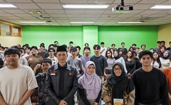 Ketua Bawaslu Provinsi DKI Jakarta berdiri di barisan depan bersama beberapa staf, berfoto dengan mahasiswa Binus yang memenuhi ruangan kelas, dengan latar dinding hijau dan perangkat proyektor di atas